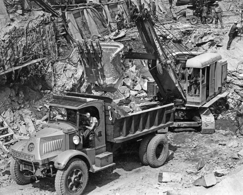 Edgar Browning Collection photo.
A Woodcrest-Rosoff Lorain shovel loads blasted rock into a Mack AC “Bulldog” heavy single axle chain drive dump truck. The truck is owned by the Andrew Gull Corporation of Brooklyn, N.Y. The work depicted is at the East River Tunnel approaches at 1st Avenue and 37th Street in New York City, circa 1937. Wagon drills hammer blast holes on the ledge above.