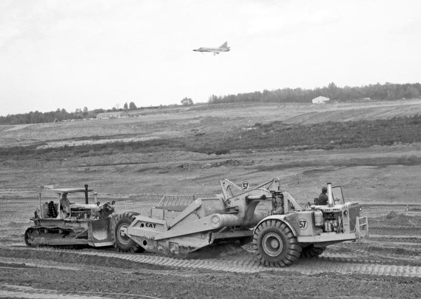 A Caterpillar DW-21 scraper being push loaded with a Caterpillar D8 bulldozer. A fighter jet soars above the AFB.