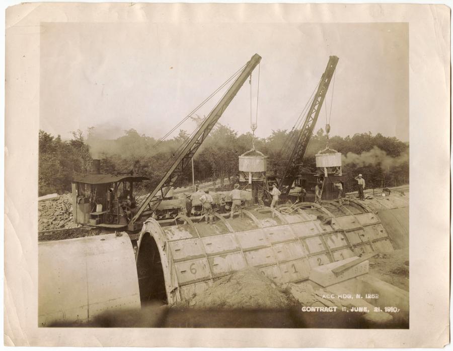 The gang is placing concrete in steel forms with revolving cranes to build a “cut-and-cover” part of the Catskill Aqueduct. Outer forms include small sections that are added in successive rows as concrete rises on each side of the arch; inside there’s a car for collapsing and moving inner steel forms. At lower right is the entrance to a siphon culvert beneath the aqueduct. June 21, 1910.
(NYC Water photo)